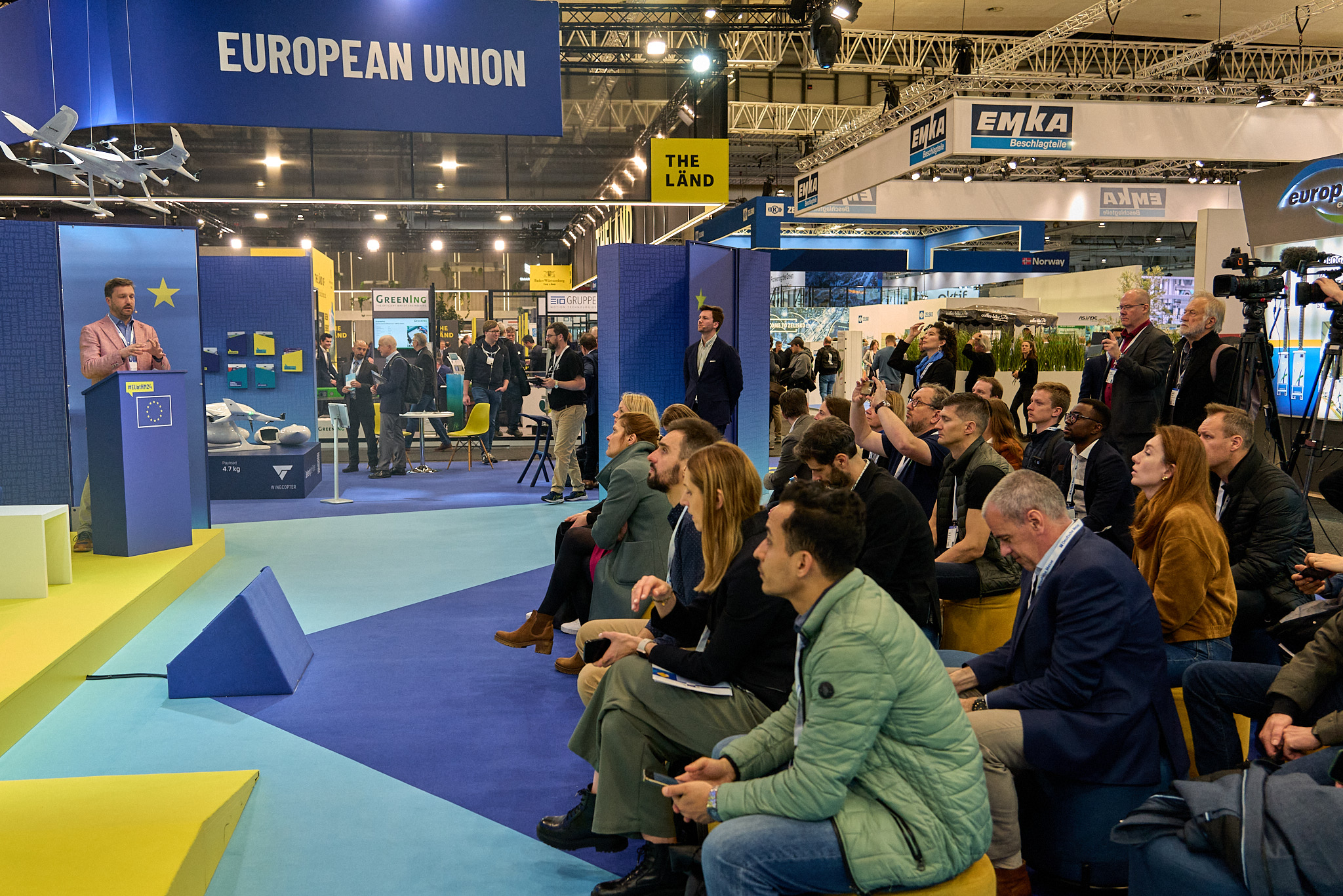 Person standing behind a lectern on stage with an engaged seated audience at the European Commission stand