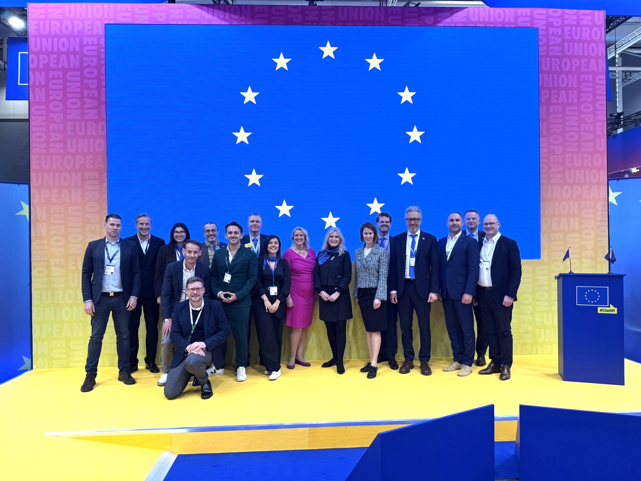 The DG GROW organising team at Hannover Messe poses for a photo on a stage. The EU flag can be seen on a screen behind them.